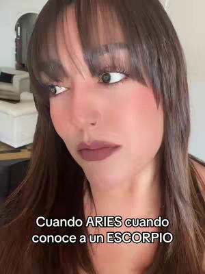 A close-up shot highlights the creator's face, featuring dark hair with bangs, rosy cheeks and a mauve lip color. The text overlay 'Cuando ARIES cuando conoce a un ESCORPIO' is placed at the bottom of the frame.