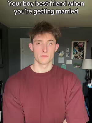 A young man in a maroon shirt poses in front of a gray wall, looking directly at the camera. The image is well-lit, and the background includes a door, a framed picture, and some wall decorations.