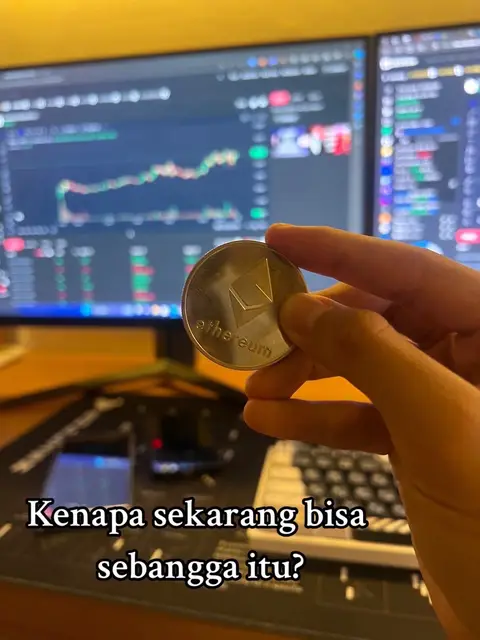 A person's hand holding an Ethereum coin is in focus, with a blurred background showing two computer monitors displaying financial charts. The hand is fair-skinned, and the coin is silver with the Ethereum logo visible. The monitors show lines and data indicative of stock market or cryptocurrency trading. The lower portion of the image includes a keyboard and a dark mousepad, suggesting a workspace. Overlayed text in the bottom left corner asks a question in a foreign language.