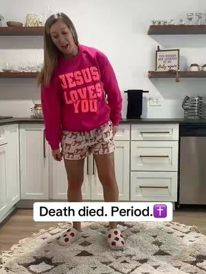 A woman stands in a kitchen, wearing a pink sweatshirt that says "JESUS LOVES YOU" in large letters. She also wears patterned shorts and fuzzy slippers. Below her is a text overlay that says, "Death died. Period." with a cross emoji.