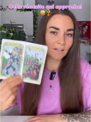 A woman is holding two tarot cards, smiling and looking at the camera. She has long brown hair and a purple shirt, with a microphone and a necklace in the front. In the background, is a kitchen setting with plants.