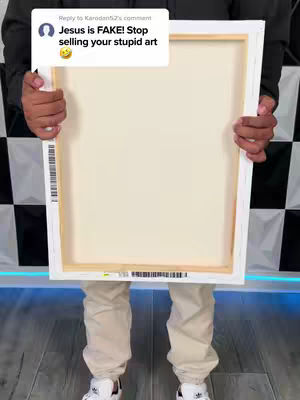 A person is holding a white canvas. The canvas is rectangular with visible wooden frame. The person is standing in front of a black and white patterned background. They are wearing a dark jacket, light-colored pants and white shoes. A comment overlay is in the top-left.