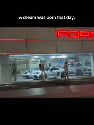 A nighttime scene outside a Porsche dealership. The focus is on the storefront, brightly lit with the Porsche logo visible. Several luxury cars are on display inside. A person stands outside the store, seemingly looking at the cars.