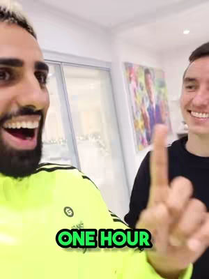 A selfie-style shot with two men; one is smiling widely, showing teeth, and wearing a bright yellow top, with a highlighted area on his blonde hair. Text "ONE HOUR" in a bold green overlay. The other man points upwards, and has a neutral expression. There is a colorful artwork and a door visible in the background.