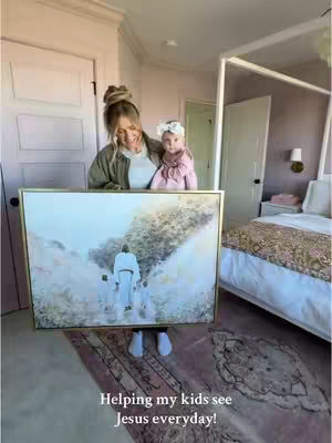 A woman with blonde hair, wearing a green jacket and a white top, holds a framed painting of Jesus with two children in front of her. She is also holding a baby. The painting rests on her forearms. The background reveals a bedroom setting with a bed, side table, and pink walls.