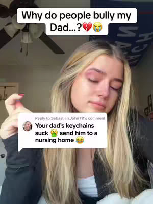 A young woman with long blonde hair is the central figure, her face expressing sadness. Above her is a text overlay asking why people bully someone's dad, with emojis indicating a broken heart and tears. Below, a comment bubble contains a reply, using emojis to express disgust and humor suggesting the dad should be sent to a nursing home. The scene is set indoors against a plain backdrop.
