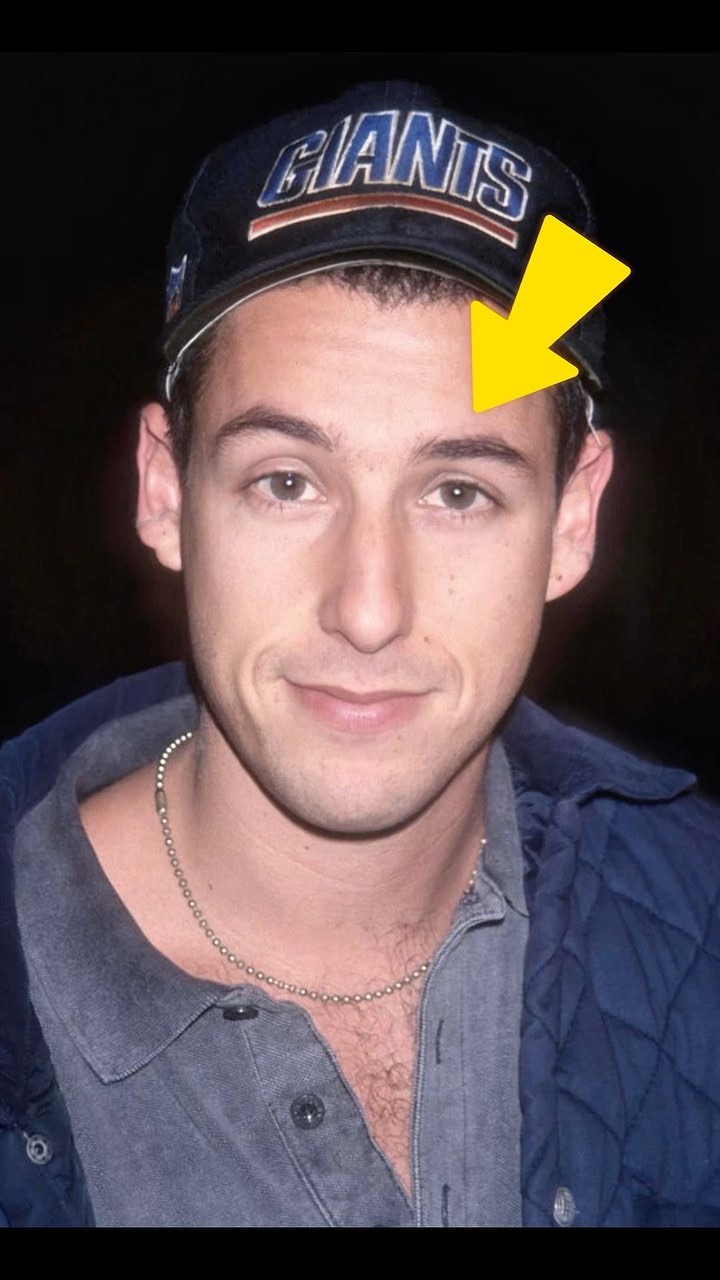 A close-up of Adam Sandler's face, smiling slightly, with a blue New York Giants baseball cap. A yellow arrow is pointing to his eyebrow. He's wearing a gray shirt and a silver chain. The background is dark.