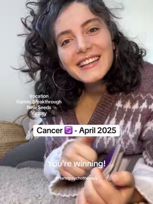 A woman with curly dark hair smiles and holds up a stack of tarot cards. She is wearing a patterned sweater, and text overlays provide information about Cancer and April 2025, along with astrological keywords like "Vocation," "Karmic Breakthrough," and "Victory."