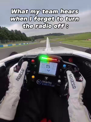 The image captures a first-person perspective from inside a race car, focusing on the steering wheel and dashboard. Text overlays the top, stating "What my team hears when I forget to turn the radio off :", framed against a backdrop of a racetrack. Dominant colors include black, white, green and the landscape of the track in the background.