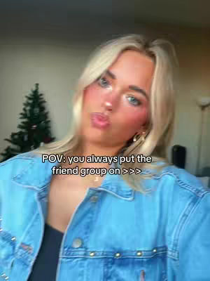 A selfie-style shot featuring a young woman with blonde hair and light makeup. She's wearing a denim jacket and gazing at the camera with a slightly puckered expression. On her face are artificial freckles. The background hints at an indoor setting, with a Christmas tree visible in the distance. Text overlay is present, starting with “POV:”.