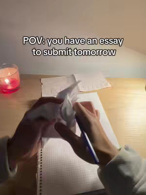 POV shot of a person holding a crumpled tissue and pen over a notebook. The scene is lit by soft desk lighting, with a candle visible on the left and a stack of papers on the desk.