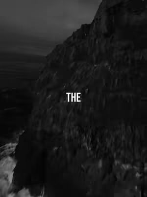 A dramatic grayscale image of a dark, rocky mountain with text overlayed. The mountain is the dominant element, filling most of the frame. The text 'THE' is centered in the lower portion of the image against a blurred background. The overall composition creates a sense of depth, with the mountain's texture and the play of light and shadow adding to the visual interest.