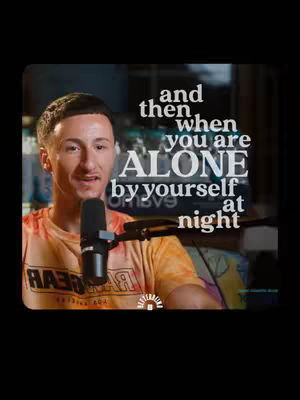 A man with short dark hair is centered in the frame, speaking into a microphone. He wears a yellow and orange patterned shirt. Overlayed on the image is white text in a handwritten font.