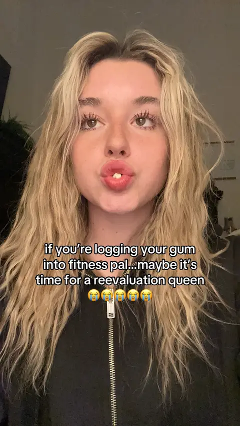 A close-up shot of a young woman with long blonde hair and full lips. She is wearing a black top and has text overlayed on top of her. The background is a muted gray.