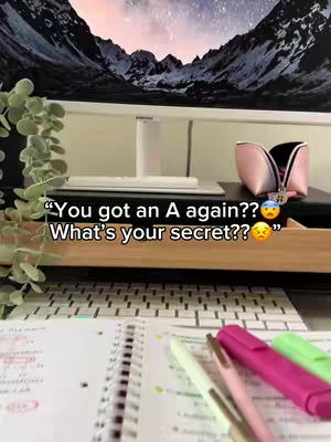 The image shows a desk setup with a computer monitor displaying a mountain landscape. In front of the monitor, there's a wooden tray holding stationary, including a book, pens, and highlighters arranged for studying. The text overlay includes the question 'What's your secret??' above the study materials.