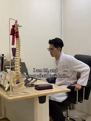 A man in a white lab coat sits next to a desk with a spine model and a laptop. Text overlay reads, "I am 23, and I'm not afraid to admit..."