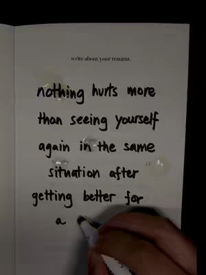 An open notebook page with handwritten text in black ink and water droplets. The text reads 'nothing hurts more than seeing yourself again in the same situation after getting better for a' The top of the page includes faint text, 'write about your trauma.' A hand holding a black marker is visible at the bottom of the image.