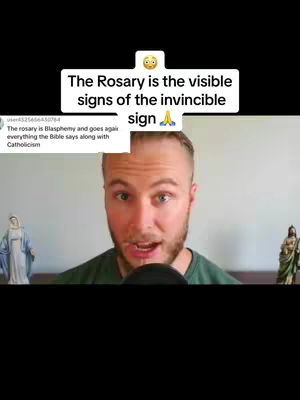 A man with a beard and a green shirt is speaking into the camera. In front of him, there's a microphone. Above him is a white banner-like graphic. On the left side of the man, behind him, there is a statue of a saint in a blue dress. On the right side, there is another statue in green.
