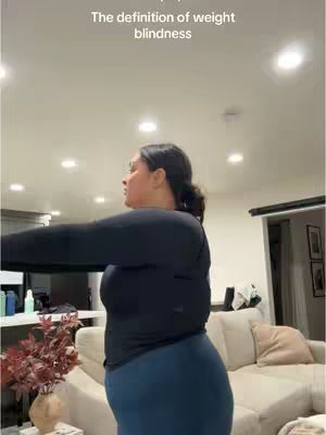 A woman in a dark blue top and leggings stands in a room, with her back to the camera but looking to the side. The background includes furniture and lighting, with text overlayed at the top.