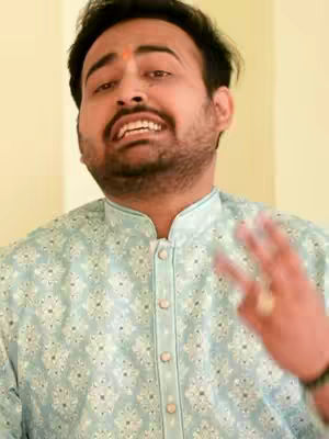 The cover image features a man with a light complexion, sporting a neatly trimmed beard, and wearing a light blue embroidered shirt. He has a red marking on his forehead. His expression is animated, with his mouth open as if he is speaking, and he is gesturing with his right hand with three fingers extended. The background is a simple, blurred, cream-colored wall.