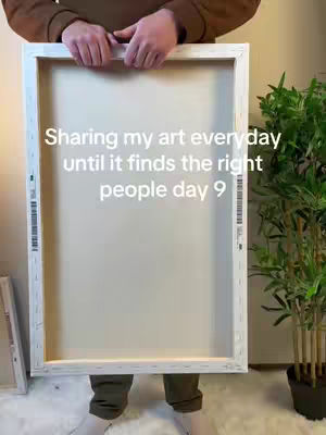 A person is holding up a large, white-framed canvas in front of their body, with text overlaid on the canvas. The person is wearing a brown long-sleeved shirt and green pants . A green plant is visible on the right side.