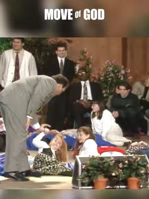 The image captures a scene from a religious service. A man in a suit leans towards a group of people lying on the floor. Other individuals, including both men and women, are standing and observing. The background suggests an auditorium setup, complemented by decorative plants.