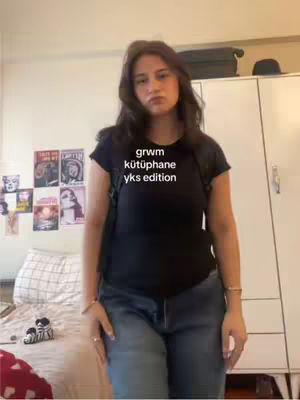 A young woman stands in her room, facing the camera. She wears a black t-shirt and jeans, with a backpack on. The room has a bed with a patterned cover, posters on the wall, and a white wardrobe. Text overlay reads: 'grwm kütüphane yks edition'.