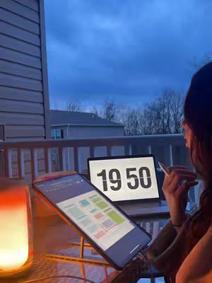 A person is working at a desk, focusing while looking at a laptop, a phone, and a glowing lamp. It is nighttime, the sky is blue, and the scene has a warm, inviting glow.