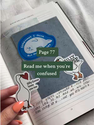 The image shows an open book with a hand holding a sticker of a goose, along with other goose and cloud stickers. Text overlays indicate the current page number and a message. The color palette is neutral with pops of blue and white from the stickers, with orange from the nail polish. The composition is framed from a downward angle.