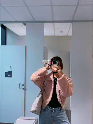 A selfie taken in a well-lit space, likely a bathroom, with a large mirror reflecting the scene. The person, likely a young woman, is holding a phone to take the picture, obscuring her face. She wears a pink fuzzy jacket, black top, and light-wash blue jeans. A pale beige tote bag with a dark pattern hangs over her shoulder. The background includes white doors, a white ceiling with recessed lighting, and a partially visible window, all reflecting in the mirror. The color scheme is soft with a focus on pinks, whites, and blues.