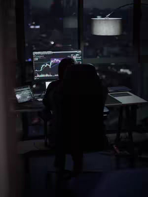 A dark, moody image of a person sitting at a desk in front of a computer screen, likely in a home office at night. The screen displays what appears to be a stock trading chart. The person is silhouetted, with the back facing the camera, and wearing dark attire. The setting suggests a focused work environment, with a city’s lights visible through the window in the background.