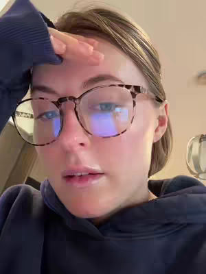 A close-up shot of a woman with fair skin and light brown hair, wearing glasses with a tortoise shell pattern. She has her hand on her forehead, with a slight expression of surprise or thought. The lighting appears to be from a screen or overhead, reflected in her glasses.