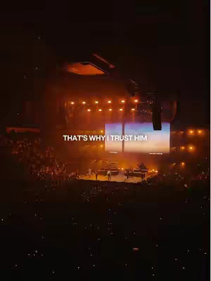 The cover image shows a concert venue at night. The foreground is filled with a crowd of people. The background features a stage with bright lights and a large screen displaying an image. Text overlayed on the image reads, 'THAT'S WHY I TRUST HIM'. The overall tone is warm with orange and yellow hues from the stage lighting.