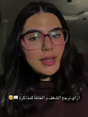 A medium shot of a young woman with dark hair and glasses, facing the camera. The background is dimly lit, with the focus on the woman's face, which is well-lit. She appears to be talking.