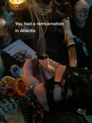 The image captures a dimly lit scene with focused elements pertaining to occult themes. In the foreground, two elegantly adorned hands are visible, displaying rings on the fingers, including what appears to be a large blue stone. The fingernails are painted black, and one finger sports a metallic finger armor. Beside the hands, the edge of a tarot card is present, adding mystical context to the frame. The background is obscured by various objects, including lit candles, additional jewelry, and a decorative knife, all contributing to a ritual or ceremonial ambiance. The color palette is dominated by dark hues, creating a mood that suggests mystery and intrigue.