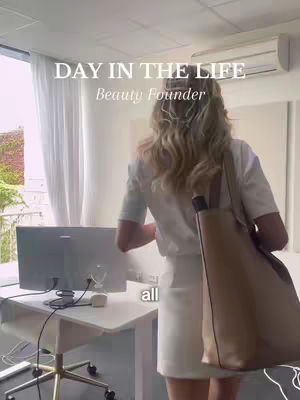 The image shows a woman from the back, standing in a modern office setting. She is wearing a white t-shirt and a white skirt, carrying a large beige tote bag over her shoulder. In the background, there is a computer on a desk, a window with white curtains, and exterior greenery visible. The text overlay reads: 'DAY IN THE LIFE' and 'Beauty Founder'.