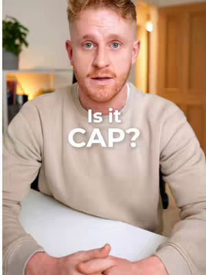 A man in a beige sweater sits in front of a white desk, with text overlayed that says, "Is it CAP?" The background is blurred, showing shelves and a plant.