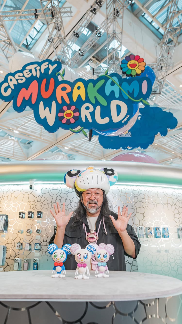 The cover image features Japanese artist Takashi Murakami in front of a pop-up installation. He is smiling and gesturing with both hands. Above him is a large sign saying 'MURAKAMI WORLD' in colorful, playful lettering, along with a smiling flower logo. Small figurines of animated characters are arranged on the counter in front of him against a backdrop of thematic floral repeating pattern.