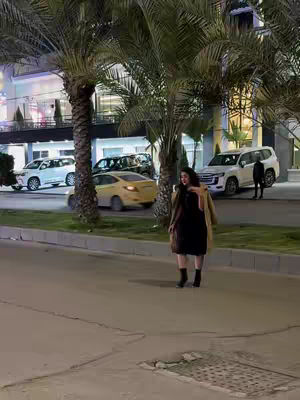 A woman in a black dress and tan coat walks along a sidewalk. Tall palm trees frame the shot, and cars are visible in the background.