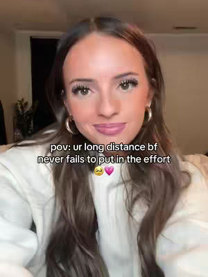 A woman with long brown hair, wearing a white sweater and hoop earrings, is centered in the frame, looking towards the camera. Text overlayed on the image reads, "pov: ur long distance bf never fails to put in the effort" with a yellow face heart eyes emoji and a pink heart emoji at the end. The background is blurred, consisting of a room interior with soft lighting.