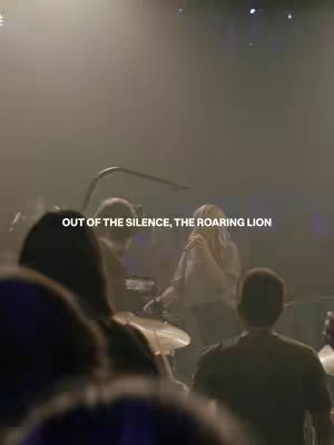 The image shows a live music performance on a stage. The focus is on a band, with a singer at the center, facing a group of people whose backs are turned towards the camera. The stage is dimly lit, and the background is blurry, filled with atmospheric haze and stage lights. The overlay text states 'OUT OF THE SILENCE, THE ROARING LION'.