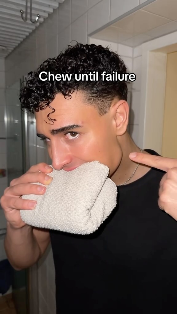 A man is seen in a bathroom setting, holding a white towel to his mouth. He's pointing with his right hand towards his jawline. The background includes a shower area and a door, suggesting a home environment. The overall composition focuses on the man's face and the towel.