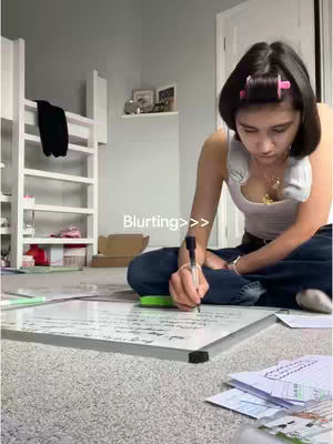 A young woman sits cross-legged on the floor in a room, writing on a whiteboard. She has rollers in her hair and is wearing a tank top and jeans. A whiteboard is in front of her with notes written on it. There is a ladder and some shelving in the background next to a white wall. The floor is covered in grey carpet.