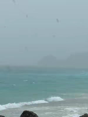 An overcast day over a seascape. The sky is a muted grey, blending seamlessly with the horizon. The ocean water is a mix of blue and green, with small waves breaking near the shore. Some dark rocks are in the foreground.