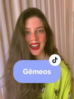 A woman with long brown hair, red lipstick, and a pearl necklace is smiling widely at the camera. A light blue rectangle with the words 'Gêmeos' is overlaid on the image. The TikTok logo is in a white circle. The background includes curtains and a small part of a lime green top. The overall composition is bright and friendly.