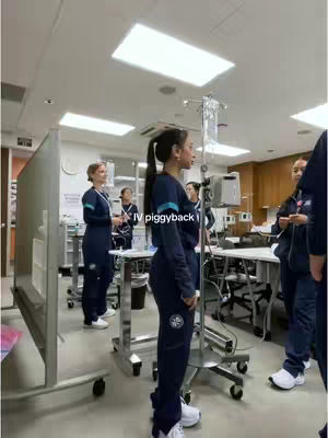 A woman in a dark blue scrub-like outfit with her hair in a ponytail is standing in a medical facility. She is the central figure, positioned in the foreground of the image, with other people in similar attire visible in the background and in the room. The lighting is bright, coming from overhead lights, and the overall setting indicates a healthcare environment, with IV stands and medical equipment.