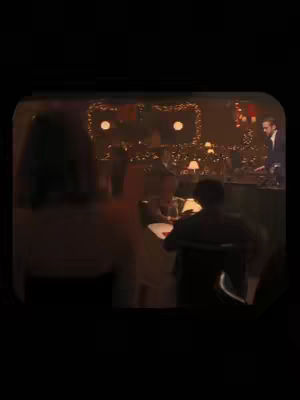 The image shows a scene inside of a dark restaurant. In the foreground, a blurred figure of a person is seated at a table. Behind them is another person. In the background, there are additional figures, possibly a bartender and other patrons, with lights above. The image is overall dark and has warm yellow tones.