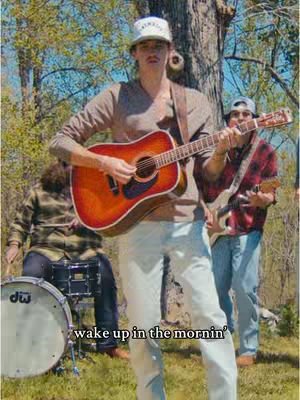 The cover image shows a band of musicians posing outdoors, with the main figure in the foreground holding an acoustic guitar. The scene is set in a grassy area with trees in the background, creating a natural and relaxed atmosphere. The color palette is warm and inviting.