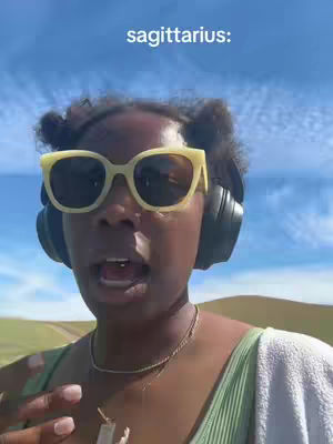 A selfie-style shot featuring a woman with her mouth open, wearing yellow sunglasses and black headphones. The sky is blue with some clouds, and the background appears to be a grassy field. Above the woman's head, the text "sagittarius:" is written in white.