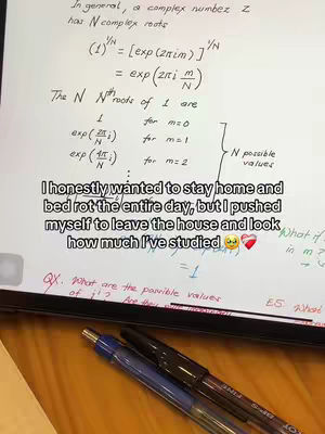 A photo taken from a student's perspective, showcasing a desk with study materials. On the left side, a tablet displays mathematical equations and handwritten notes. Two pens, one blue and one black, are placed at the bottom, suggesting active writing. The background appears blurred, focusing attention on the study setup.